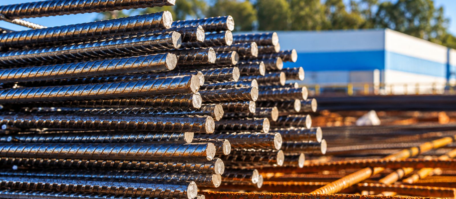 Stacked steel reinforcing bars in a yard, with a factory building behind